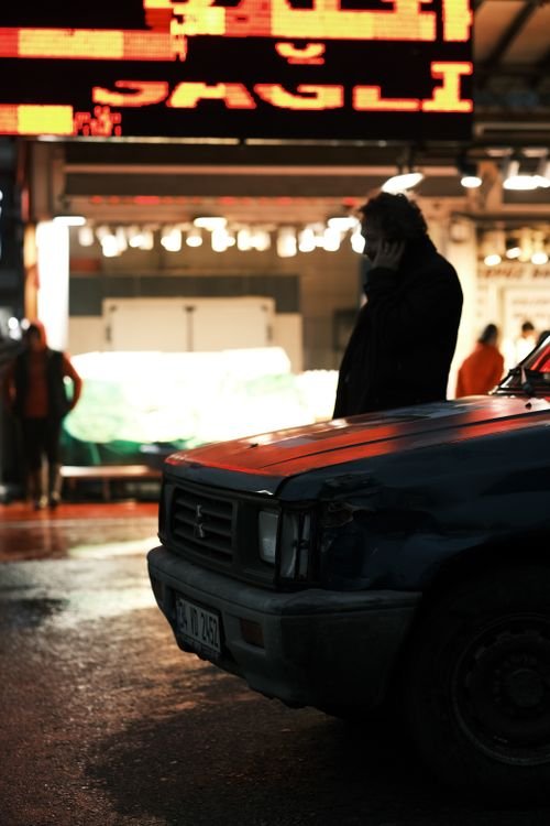 Silhouette of a man talking on a cellphone beside a parked vehicle on a rainy night city street